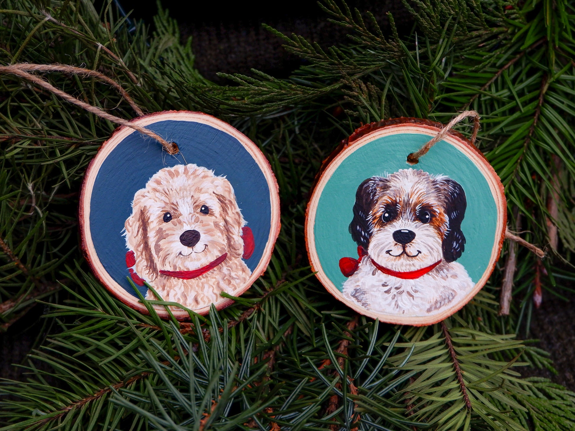 Two wooden ornament slices with dog paintings on a green pine branch background.