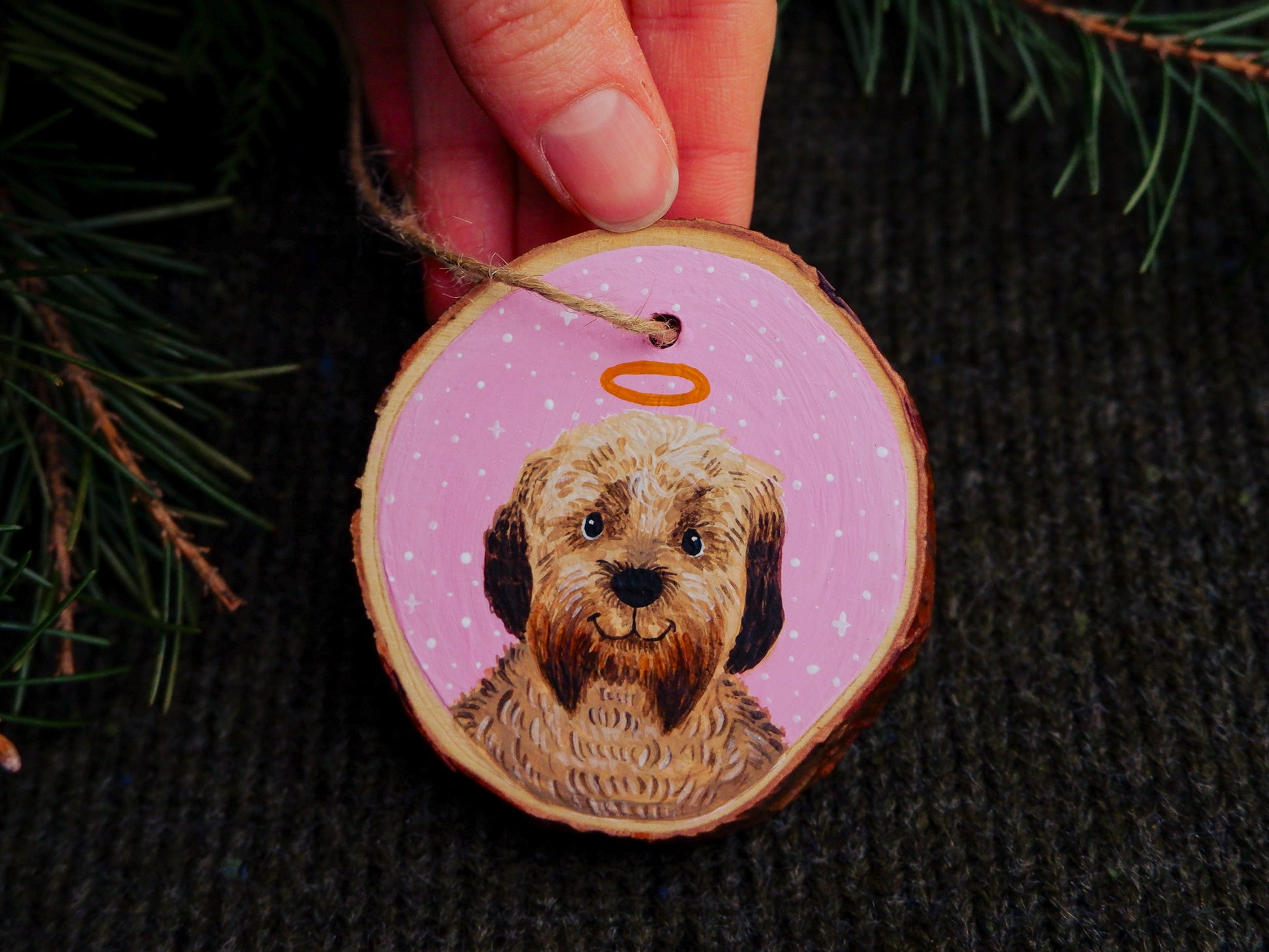 Wooden ornament with a dog painting and pink snowy background, held against a dark textured background.