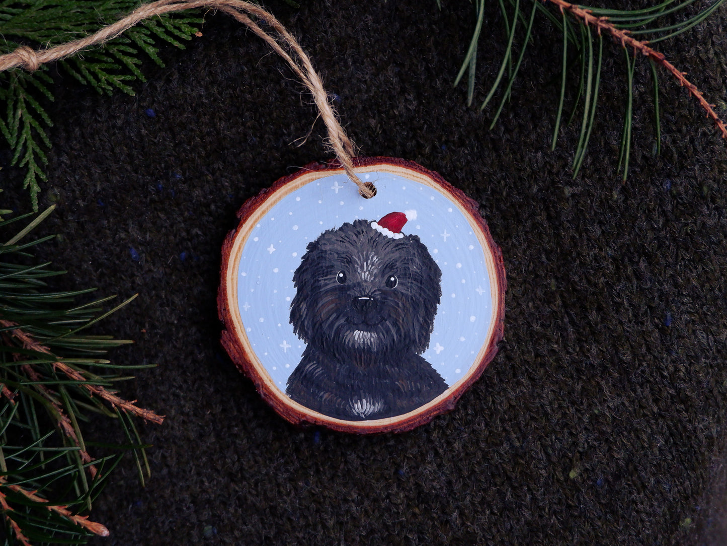 Decorative Christmas ornament with a hand-painted black dog wearing a red Santa hat on a dark fabric background.