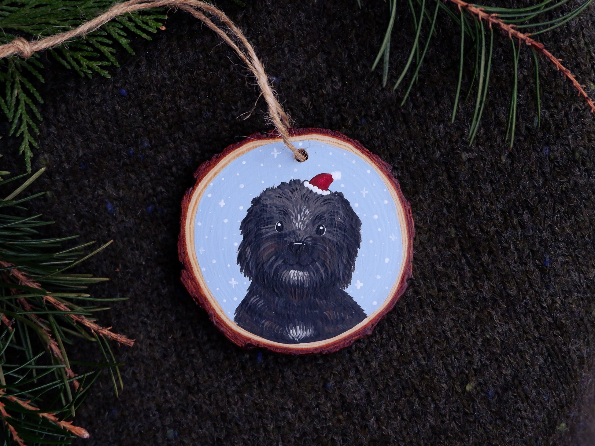 Decorative Christmas ornament with a hand-painted black dog wearing a red Santa hat on a dark fabric background.