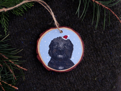 Decorative Christmas ornament with a hand-painted black dog wearing a red Santa hat on a dark fabric background.